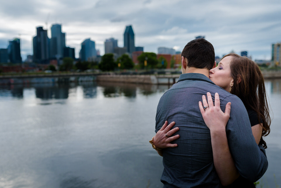 Engaged couple kissing along Lachine Canal with Montreal skyline behind