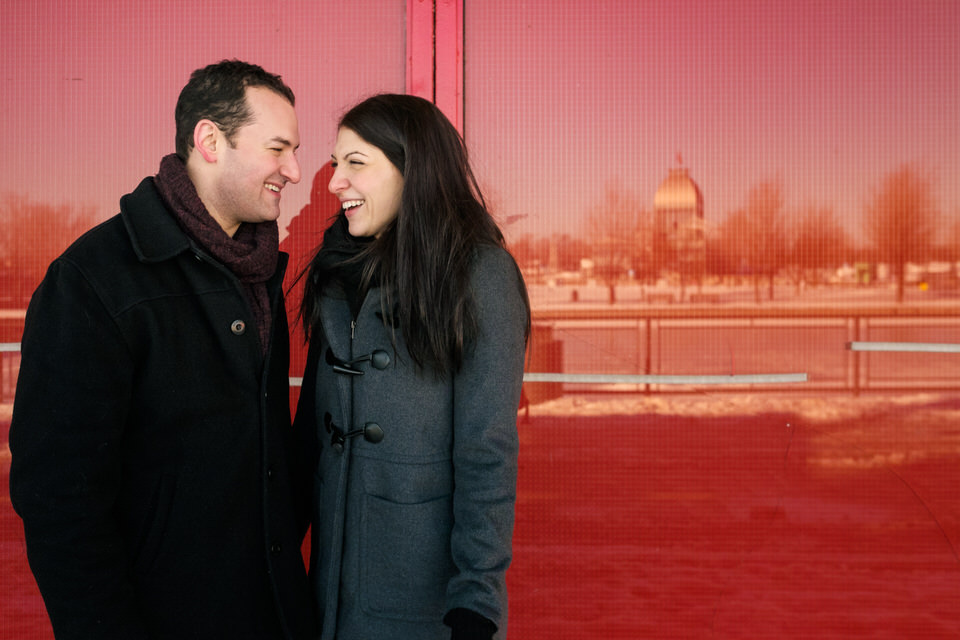 Couple smiling at each other standing next to a glass wall with the reflecion of Old Montreal Port