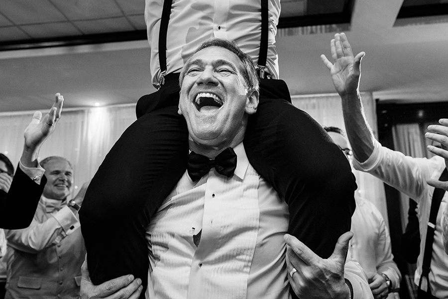 Candid photo of the father of the groom lifting his son on his shoulders, during the hora, as his shirt button pops and a huge grin covers his face