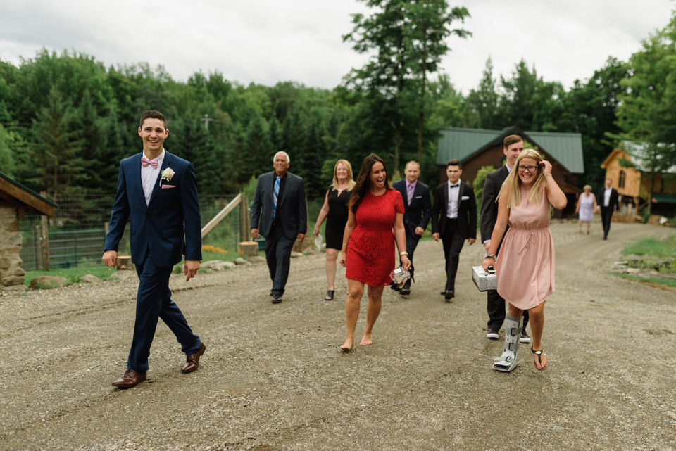 Groom walking up a gravel road with wedding guests including a woman in a boot cast