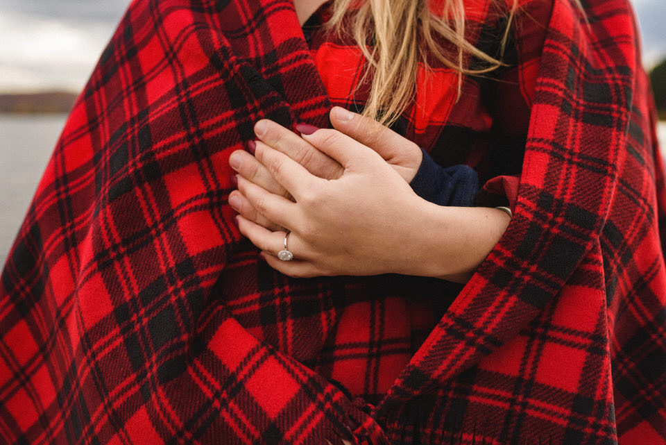Close up two hands intertwined with woman's hand with engagement ring against red and black flannel blanket