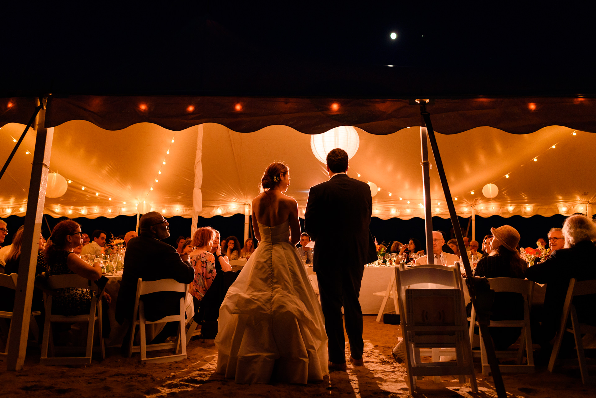 Wedding couple speaking to their guests under the warm glowing lights of a marquee tent on the beach