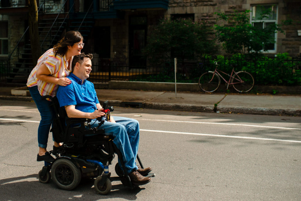 Photo of woman riding on the back of her fiancé's wheelchair as he rolls down the street