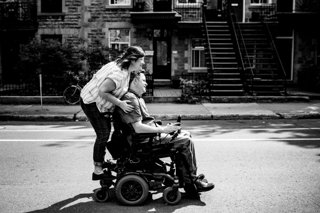 Couple rolling down the street, with woman riding on the back of wheelchair
