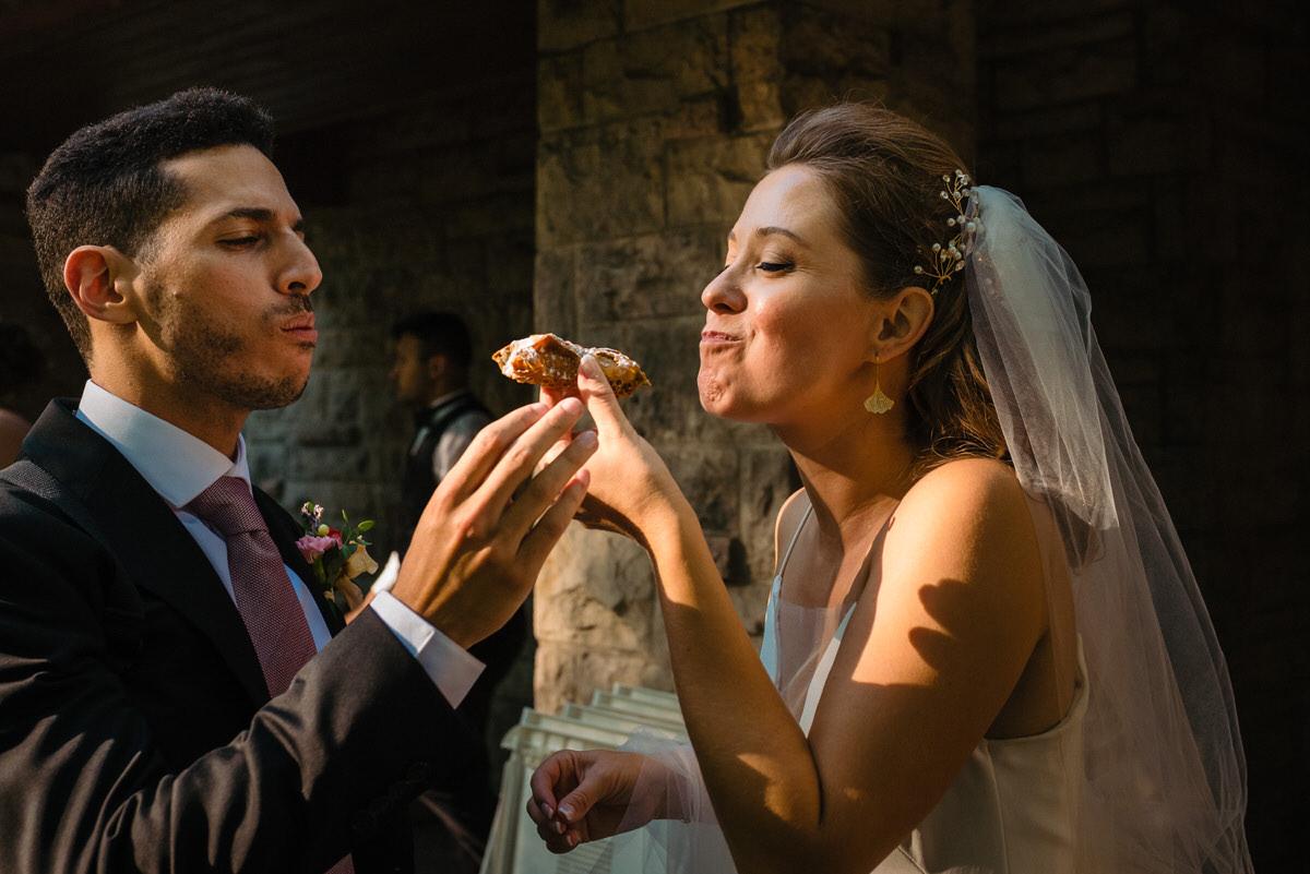 Bride and groom sharing a cannoli during cocktail hour in park