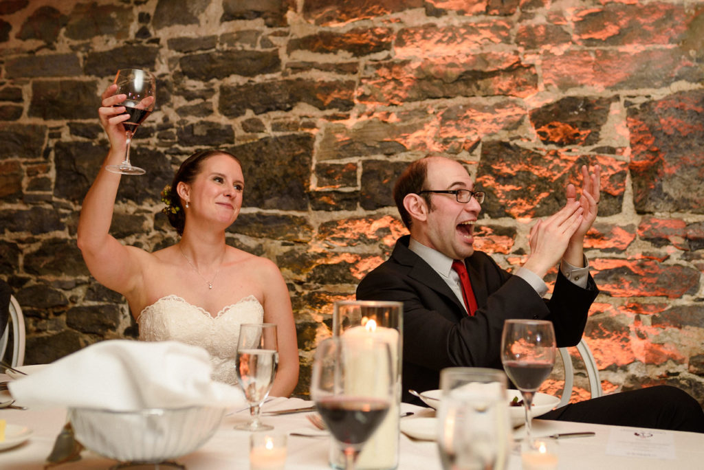 Bride raising her glass while groom laughs and claps