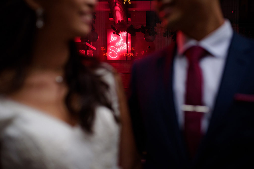 Close up on a neon sign that says "La vie est sweet" with the blurry bride and groom in foreground