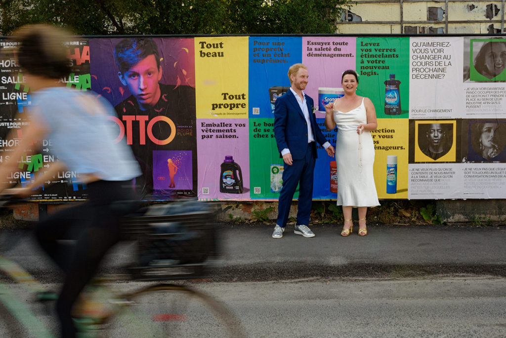 Portrait of wedding couple near a wall of posters with a biker rolling by in the foreground
